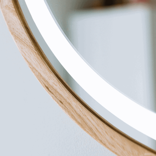 Zbliżenie na okrągłe lustro Pitagoras-Perfection-Slim-LED2.png z drewnianą ramą i podświetlanym białym obramowaniem LED, odbijające rozmyte kształty w minimalistycznym, jasnym pomieszczeniu.