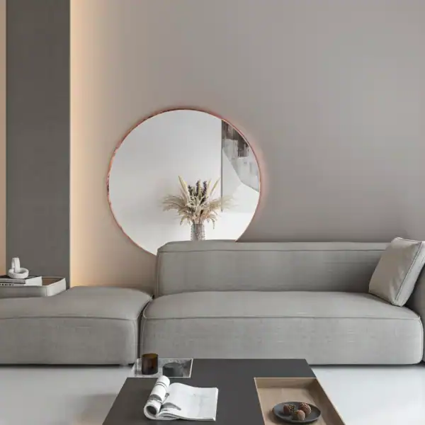 W nowoczesnym salonie znajduje się jasnoszara sofa segmentowa miedz-scaled-1.webp, okrągłe lustro ścienne i neutralny wystrój inspirowany stylem Rama. Czarny stolik kawowy z czasopismami i przekąskami uzupełniają suszone kwiaty w wazonie obok lustra.