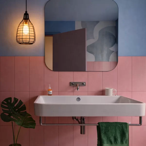 Nowoczesna łazienka z różowymi kafelkami i niebieską ścianą, z dużą białą umywalką, zielonym ręcznikiem, lampą wiszącą Kubi bez led, kwadratowym lustrem, dozownikiem mydła, kubkiem i zielonym wazonem z liściem monstery.